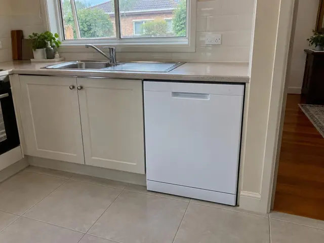 White dishwasher built into a cream kitchen beside a stainless sink and tiled splashback in Wallsend